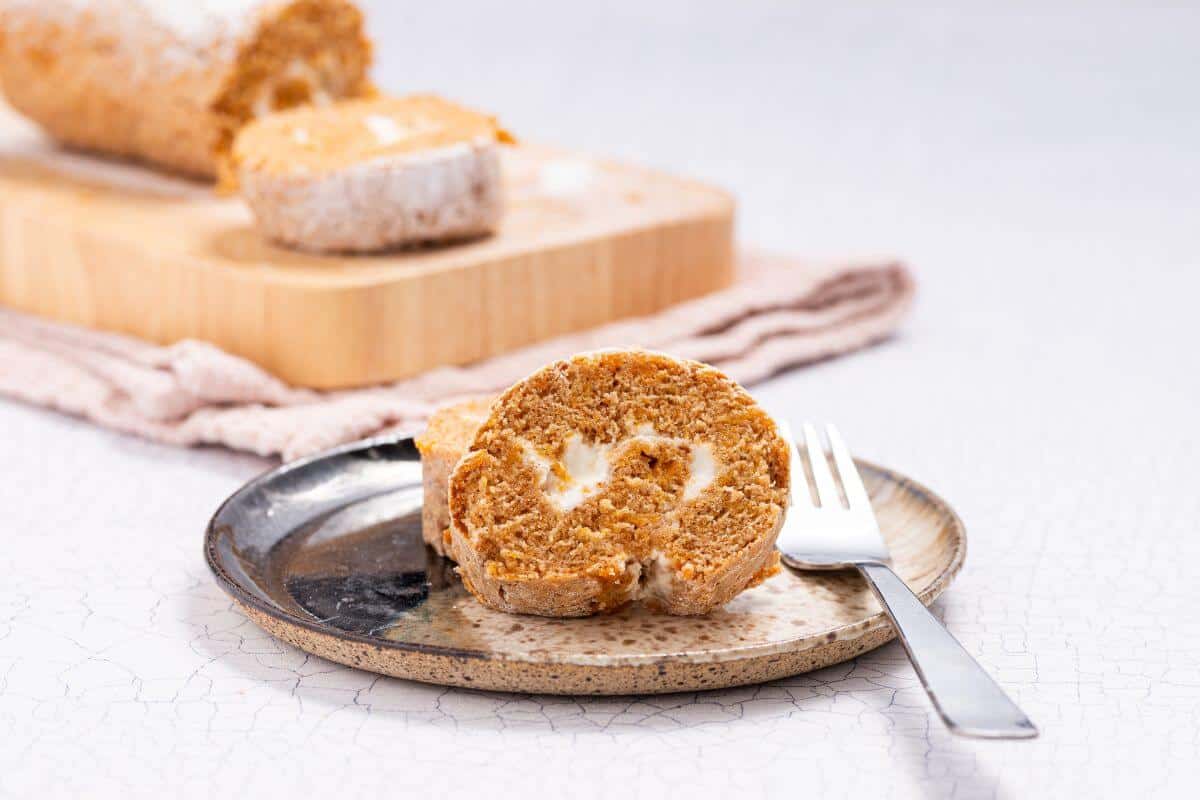 A slice of pumpkin roll cake with cream cheese filling sits on a ceramic plate next to a fork, with additional slices on a wooden board in the background.