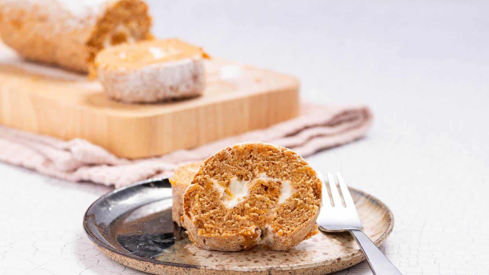 A slice of pumpkin roll cake filled with cream cheese on a plate with a fork, with the remaining cake on a cutting board in the background.