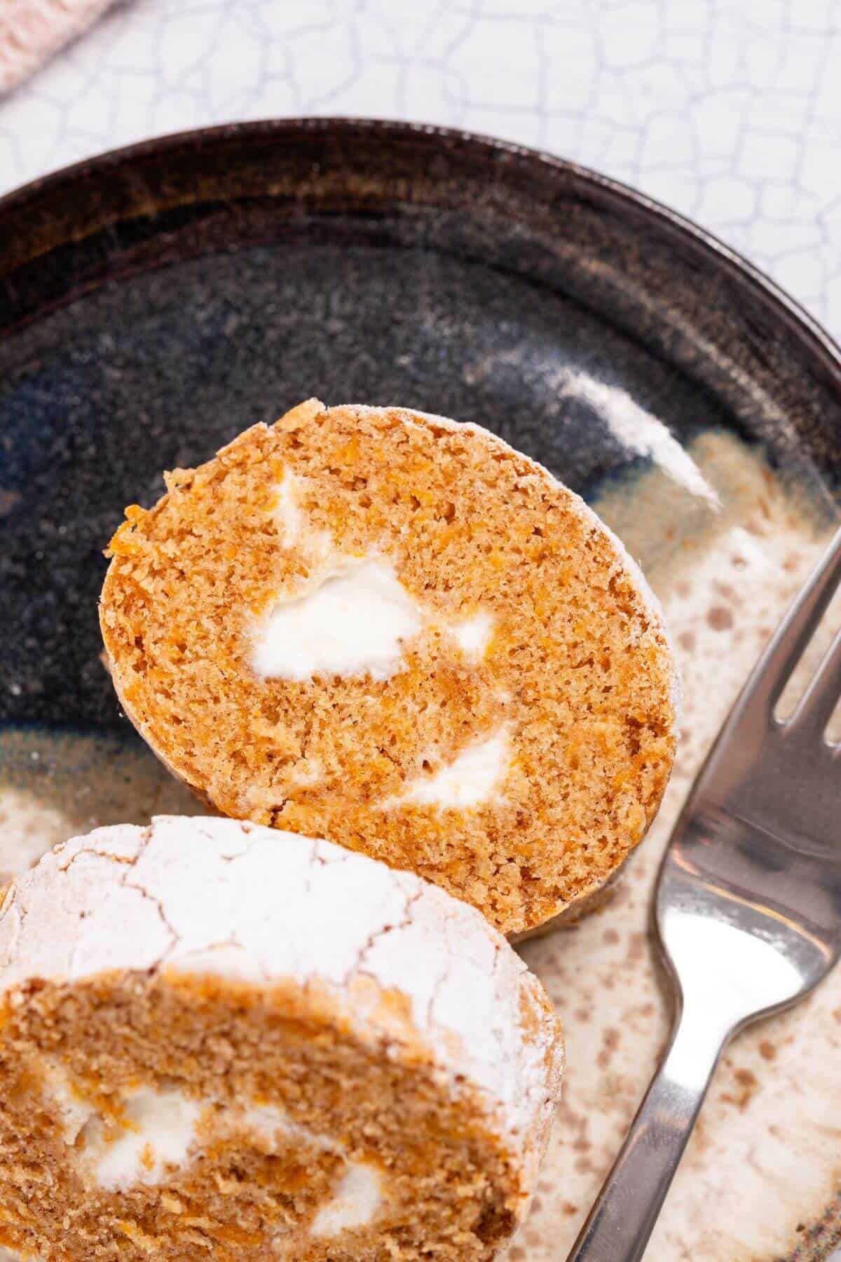 Close-up of a sliced pumpkin roll cake with white cream filling on a dark plate, accompanied by a fork.