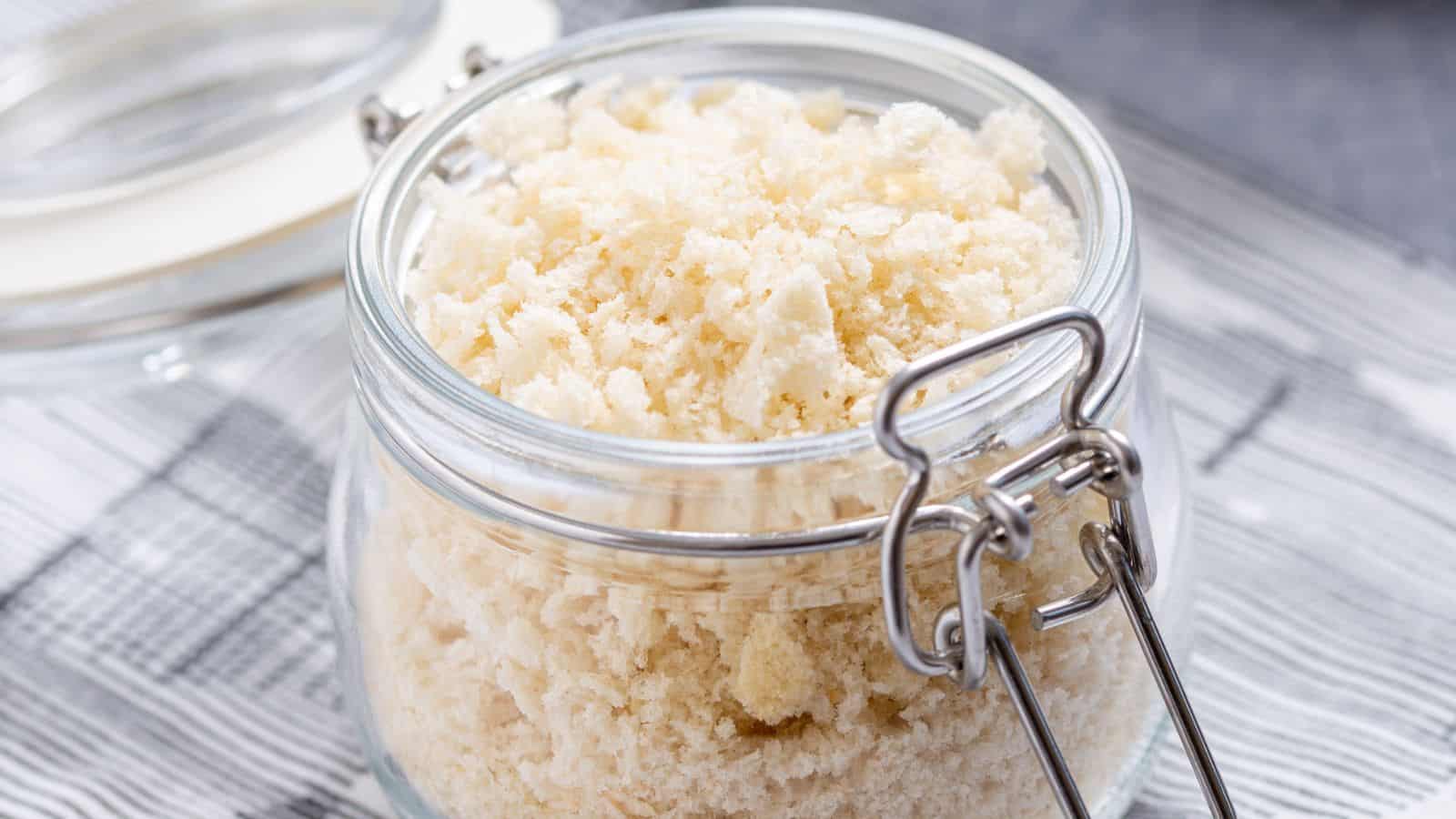 A glass jar with a metal clasp contains fluffy, off-white panko breadcrumbs, placed on a striped cloth.