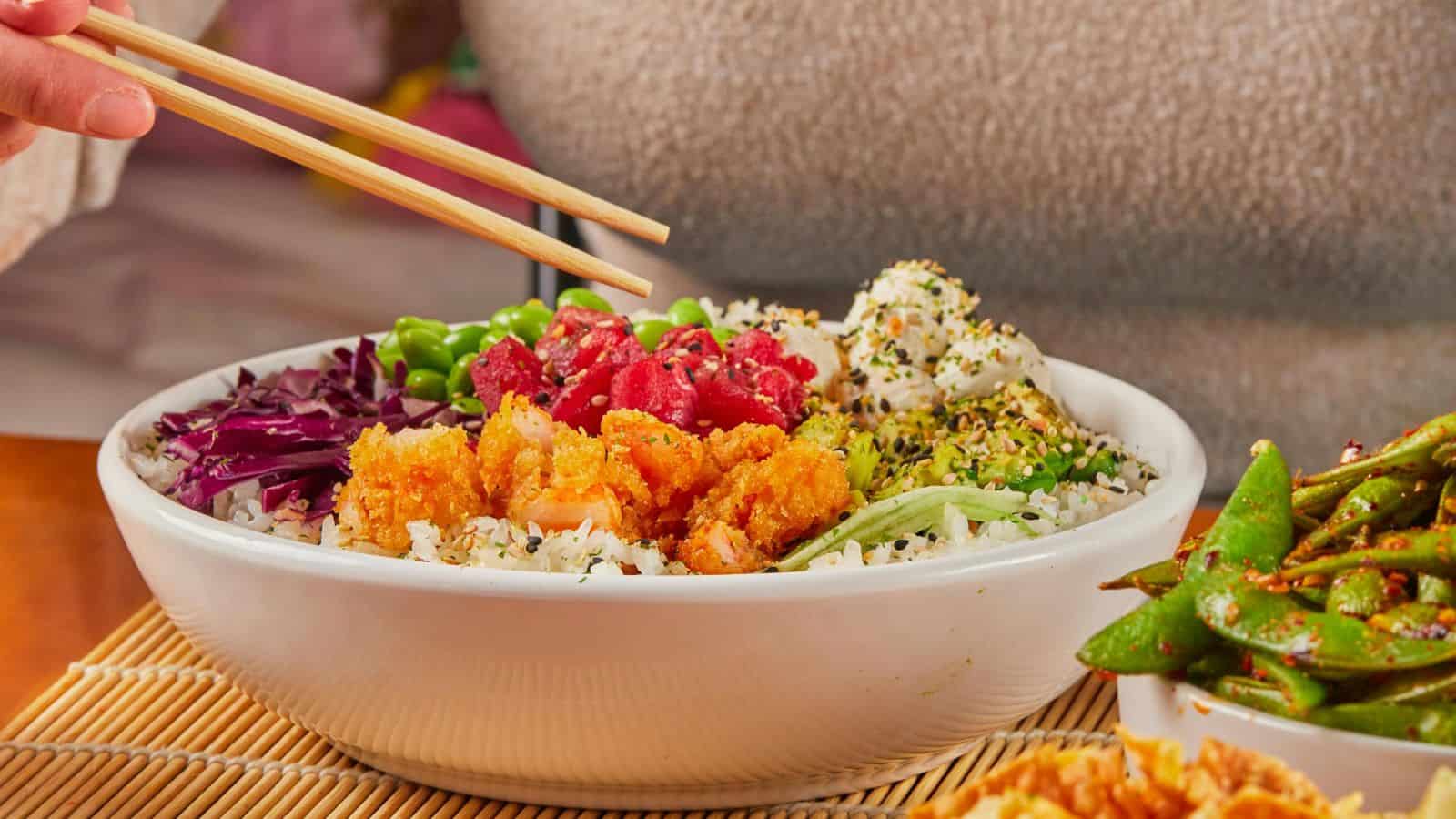 A bowl of rice topped with fish, cabbage, edamame, and seaweed seasoning, with a person using chopsticks. Edamame on the side.