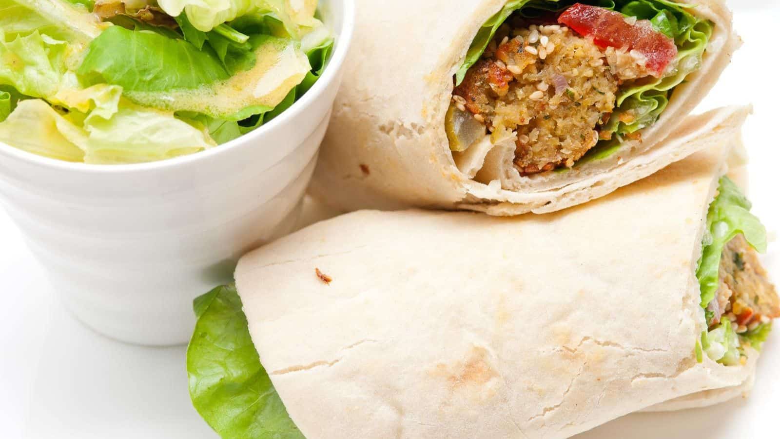 Close-up of a falafel wrap sliced in half on a plate, next to a bowl of green salad with dressing.