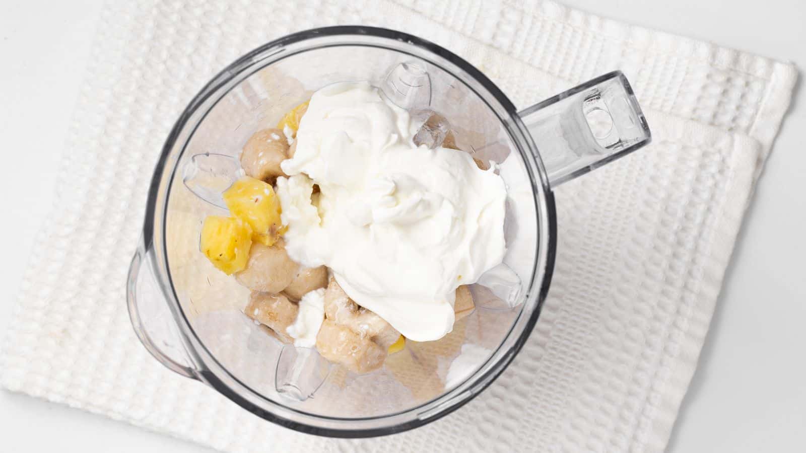 A blender with chopped banana, pineapple, and a dollop of yogurt on a white towel.