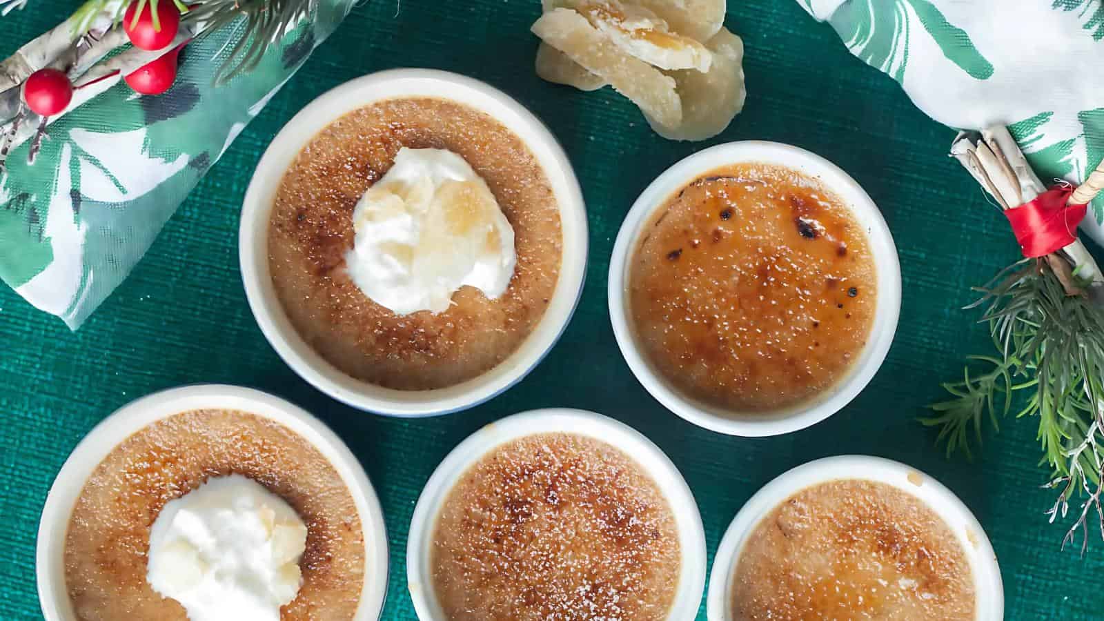 Five ramekins of crème brûlée topped with cream are arranged on a green surface, surrounded by a decorative napkin and a sprig of greenery.