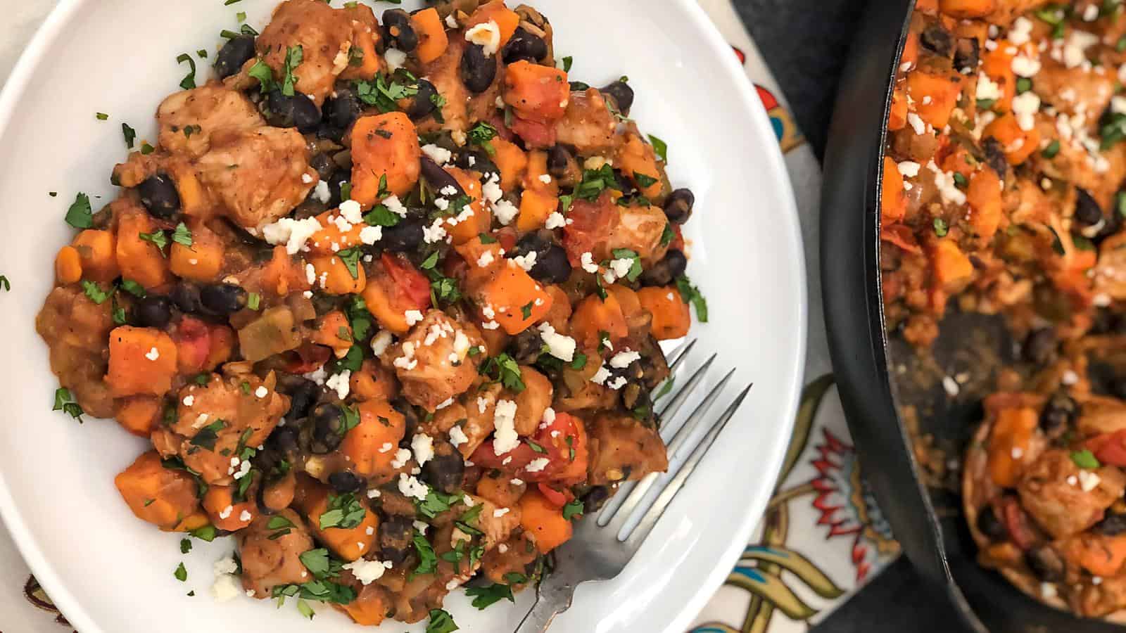 A plate of chili with chicken, black beans, sweet potatoes, tomatoes, and herbs, garnished with crumbled cheese.