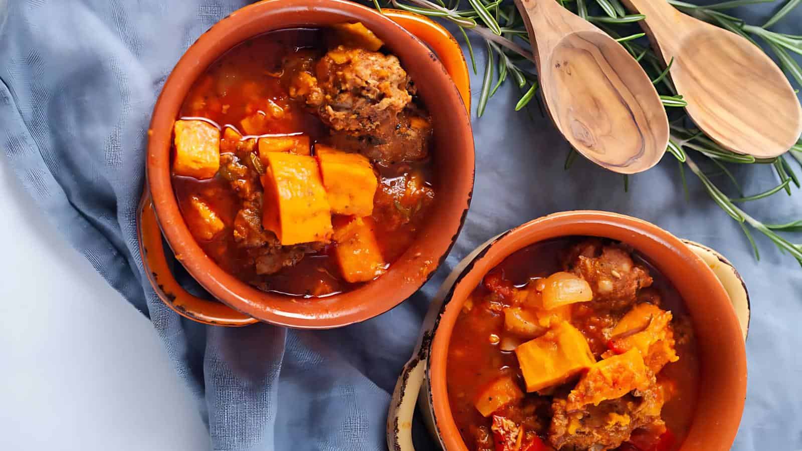 Two bowls of hearty stew with chunks of meat and vegetables, placed on a blue cloth with wooden spoons and rosemary sprigs beside them.