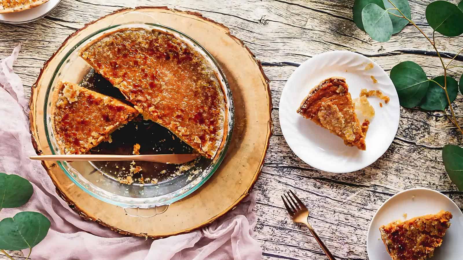 A partially sliced pie in a glass dish on a wooden board. Two slices are served on white plates. Leaves and fabric are arranged decoratively around the setup.
