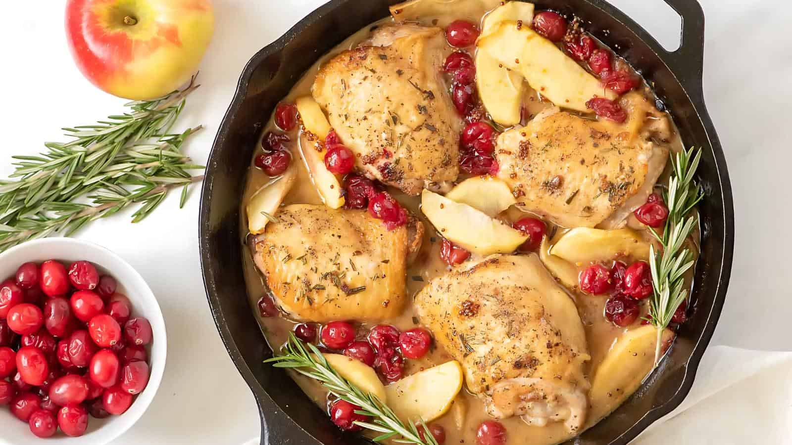 Chicken thighs with sliced apples and cranberries in a skillet, garnished with rosemary. An apple and a bowl of cranberries are beside the skillet.