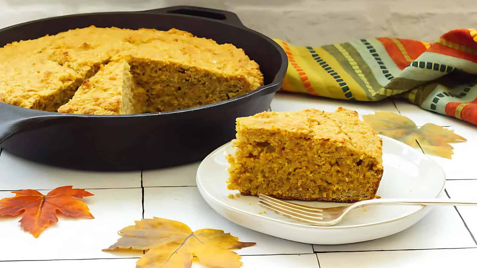 A slice of cornbread on a white plate with a fork, set beside a skillet with the remaining cornbread. Autumn leaves and a striped cloth are in the background.
