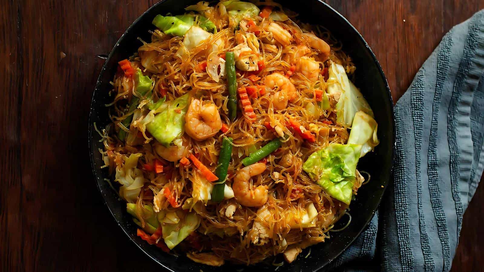 A plate of stir-fried noodles with shrimp, vegetables, and spices on a wooden table, next to a folded striped cloth.