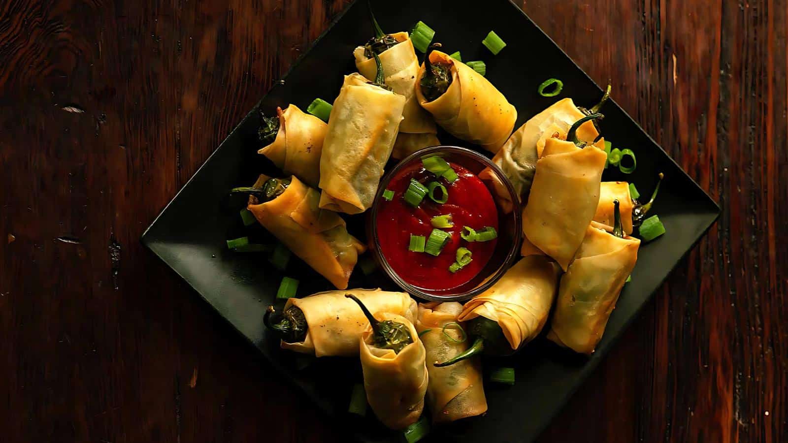 A plate of jalapeño poppers wrapped in crispy pastry, arranged around a bowl of red dipping sauce, garnished with chopped green onions on a dark wooden table.