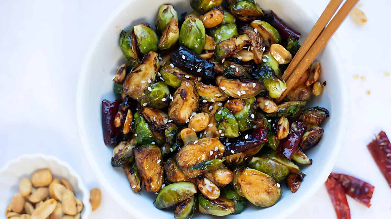 A bowl of stir-fried Brussels sprouts with peanuts, dried red chilies, and sesame seeds, with chopsticks resting on the rim.