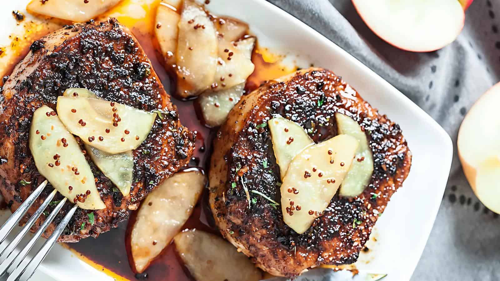 Two seasoned pork chops topped with apple slices and sauce on a white plate, garnished with herbs, surrounded by more apple slices.