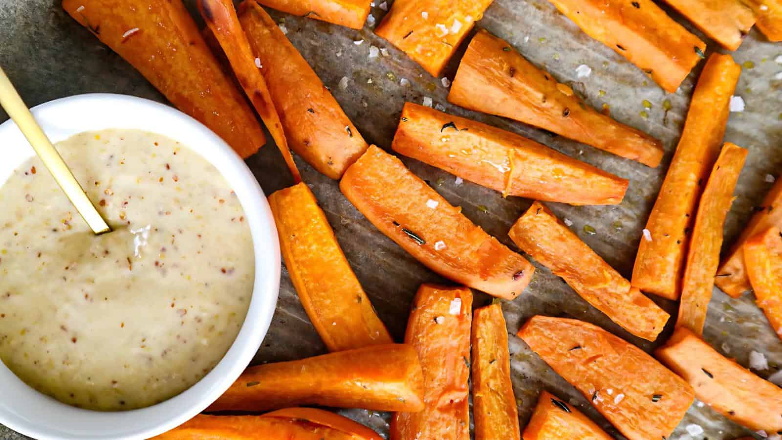 Roasted sweet potato wedges on parchment paper, sprinkled with salt, next to a bowl of creamy mustard dipping sauce with a spoon.