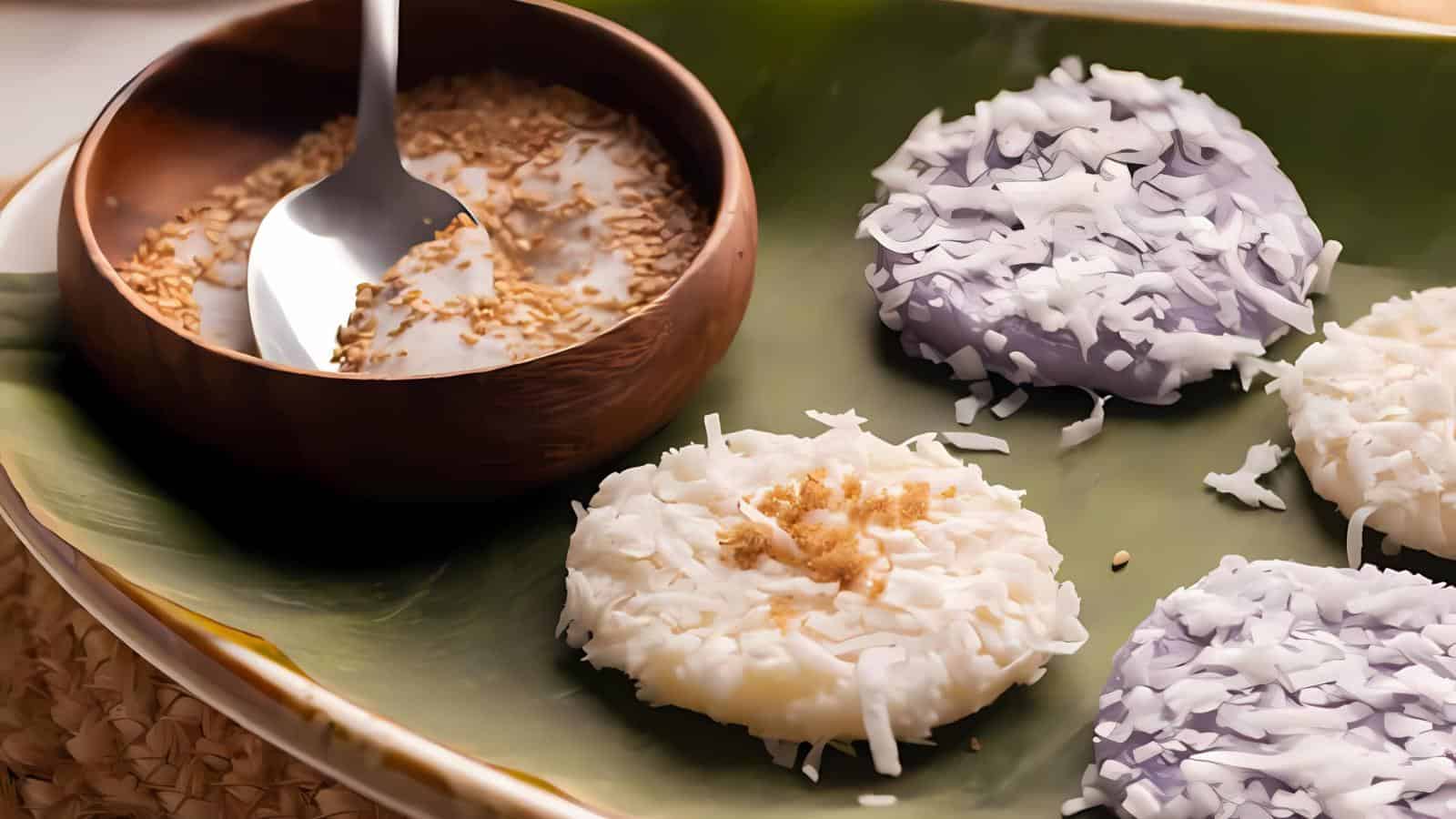 Four mochi rice cakes topped with shredded coconut on a banana leaf, accompanied by a bowl of sesame seeds and a spoon.