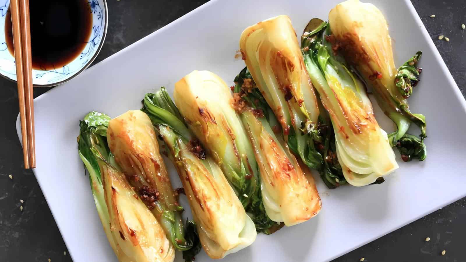 A rectangular white plate with roasted bok choy garnished with crispy bits, accompanied by a small bowl of dipping sauce and chopsticks on the side.