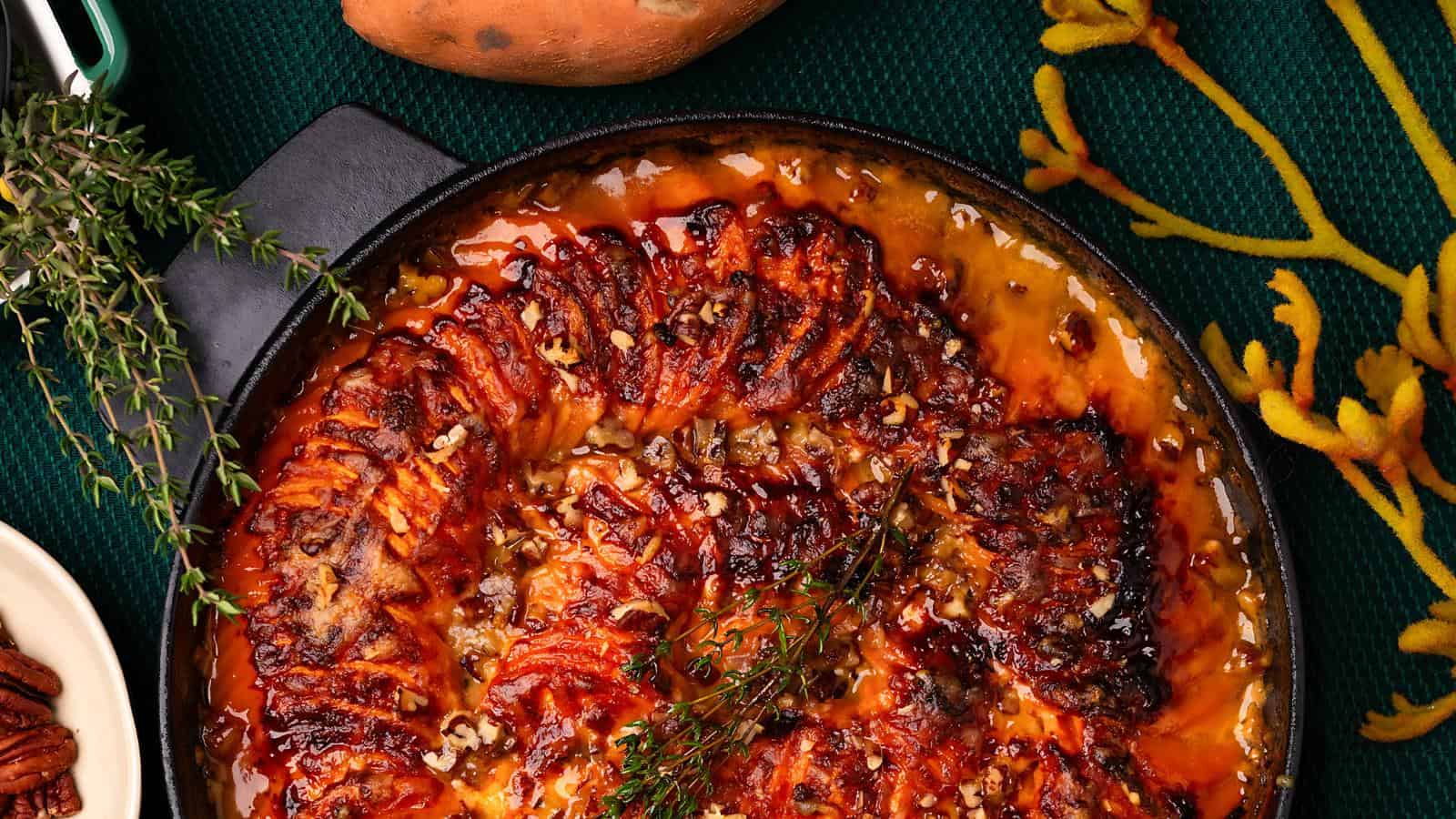 A cast iron skillet with roasted, spiral-sliced sweet potatoes glazed with caramelized sauce and garnished with sprigs of thyme, placed on a dark green surface.
