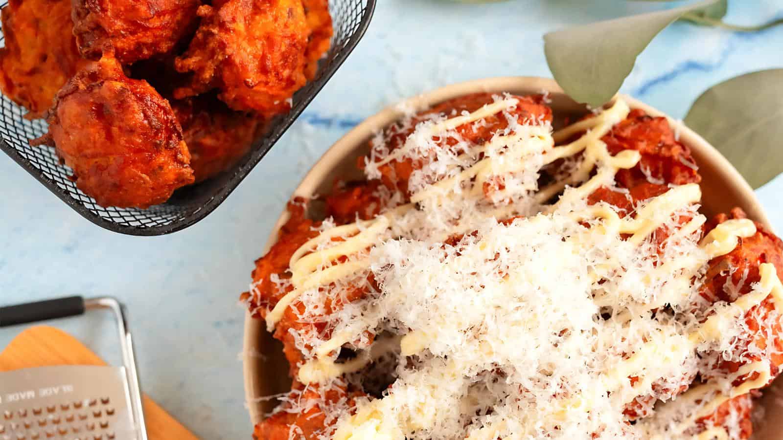 Crispy fried bites topped with grated cheese and drizzle, beside a basket of additional fried bites on a blue background.