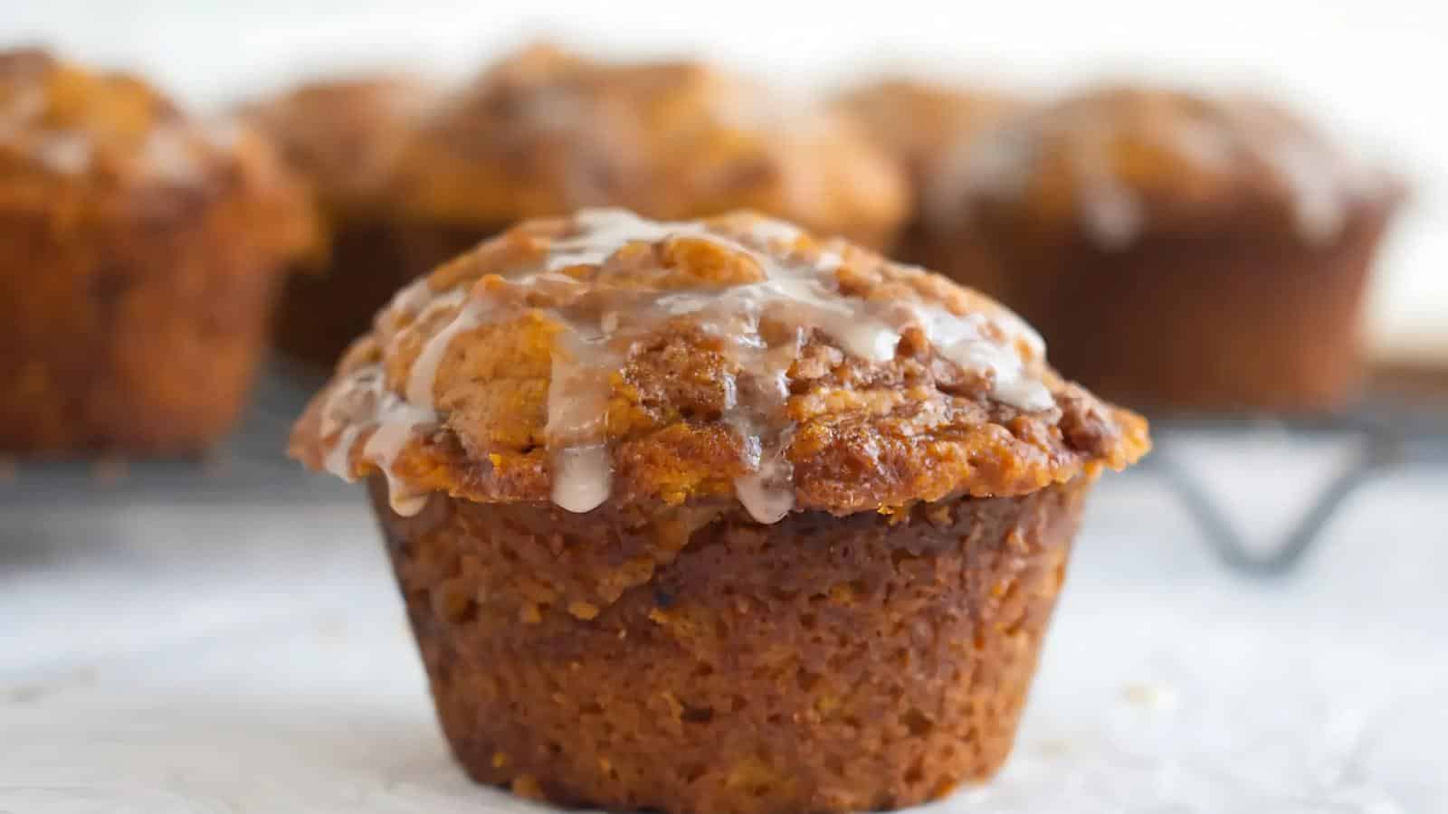 Close-up of a spiced muffin with icing drizzled on top, placed on a cooling rack with more muffins blurred in the background.