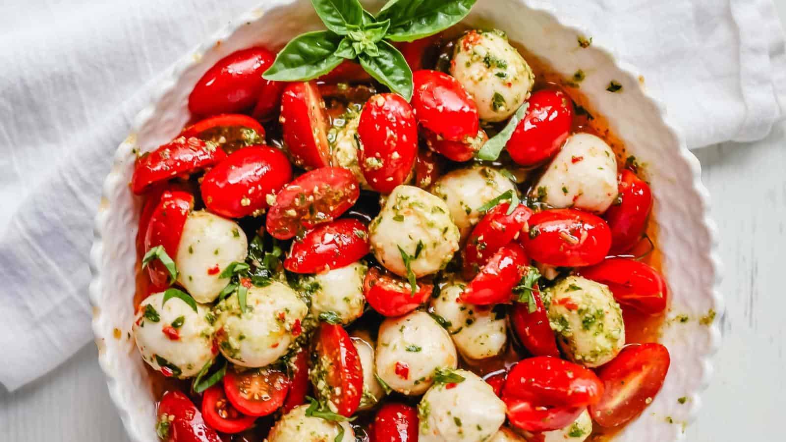 A bowl of caprese salad with cherry tomatoes, mozzarella balls, pesto, and fresh basil leaves on a white cloth.