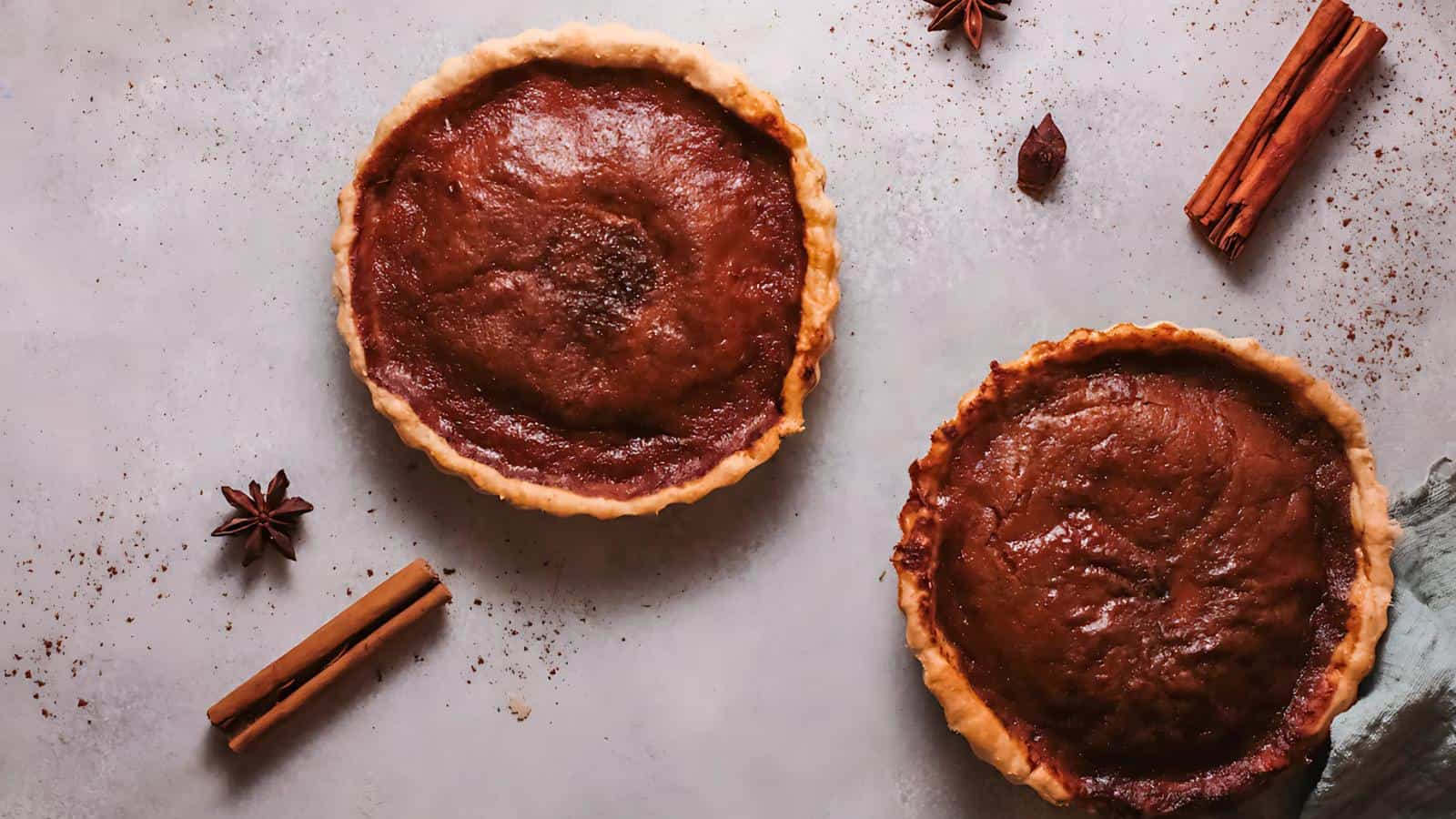 Two pumpkin pies on a gray surface, surrounded by cinnamon sticks and star anise.