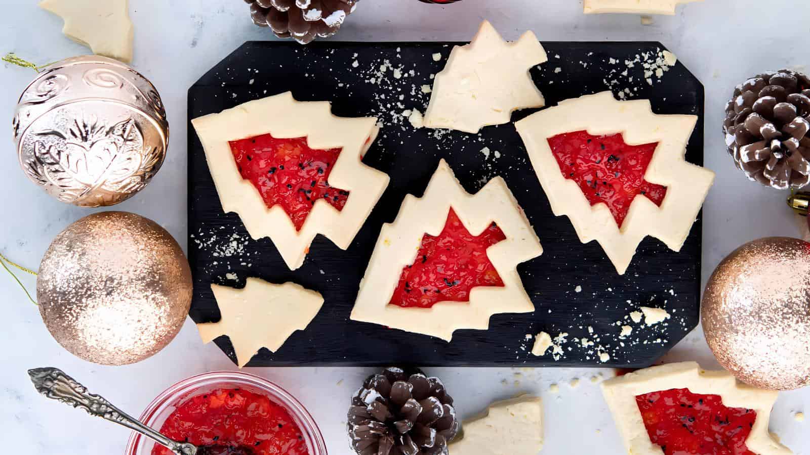 Christmas tree-shaped cookies with red filling are arranged on a black tray, surrounded by pinecones and gold ornaments.