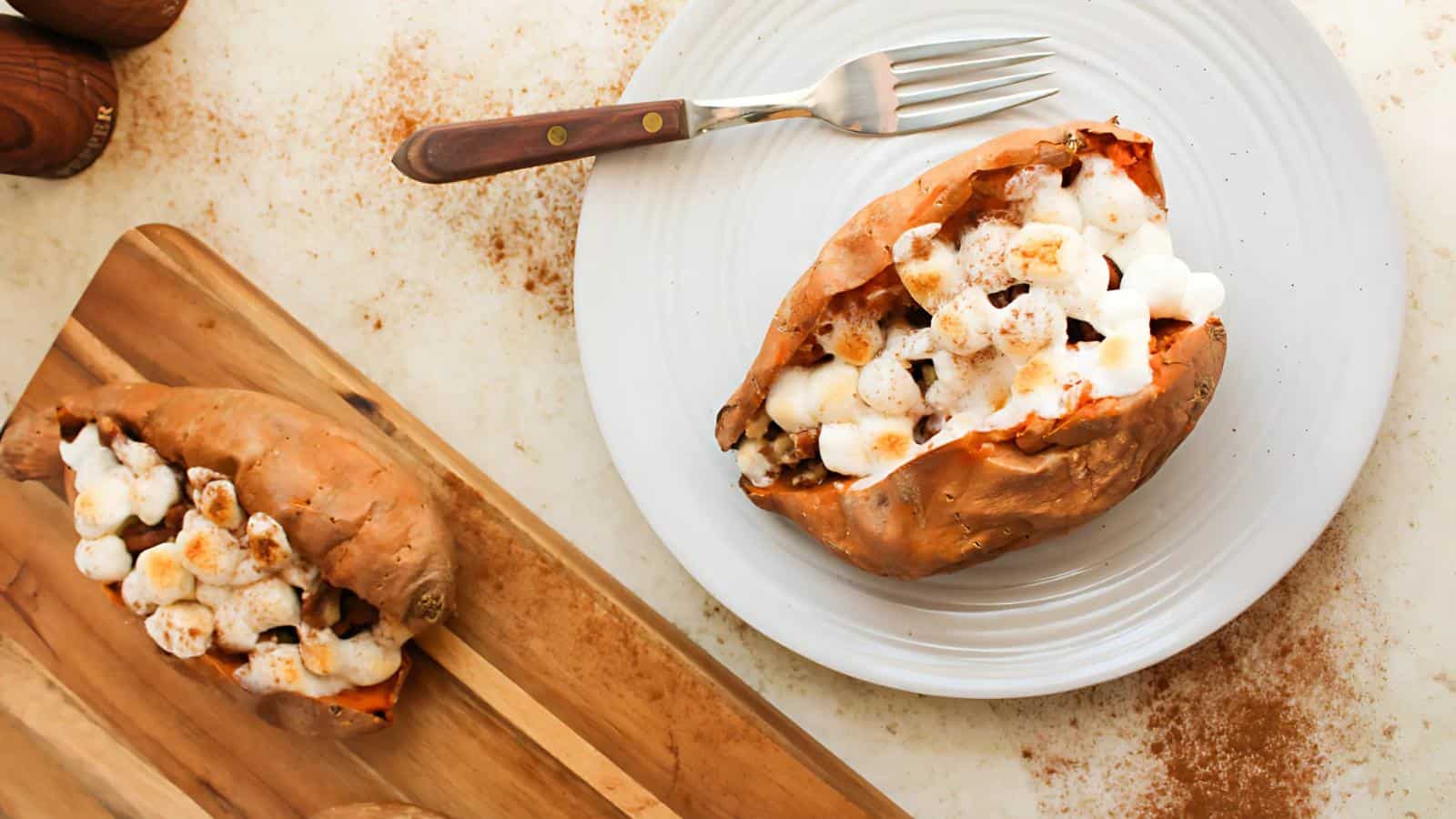 Baked sweet potato topped with marshmallows and cinnamon on a white plate, with a fork beside it. Another sweet potato is on a wooden board nearby.