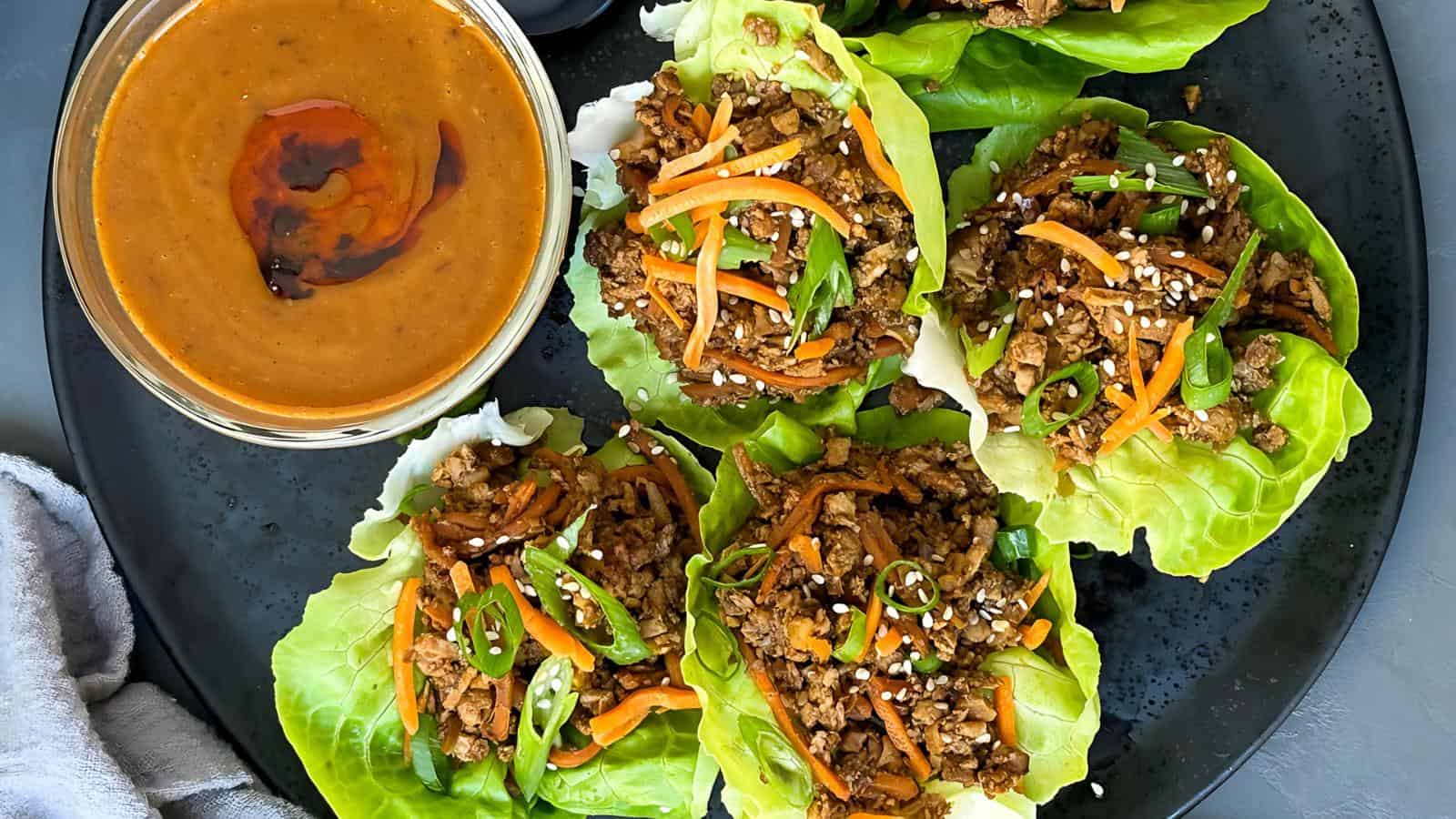 Lettuce wraps with seasoned filling and shredded carrots on a plate, served with a bowl of dipping sauce.
