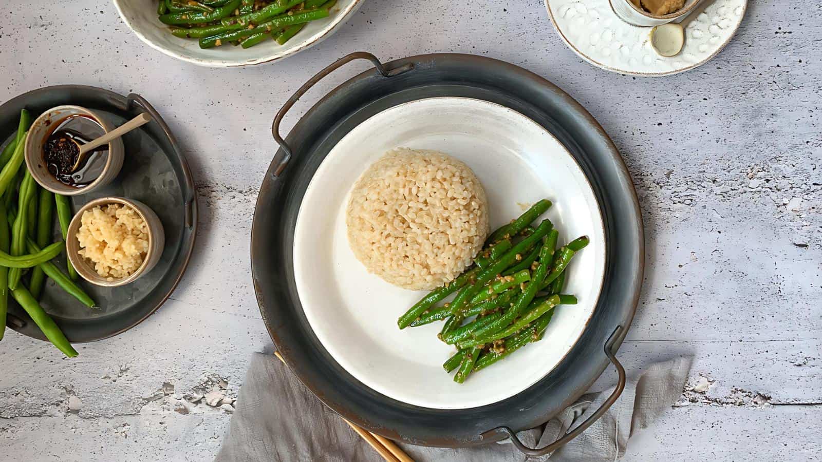 A plate with rice and green beans, surrounded by bowls of green beans, a sauce, and a grated ingredient on a textured surface. Chopsticks are placed on a napkin nearby.