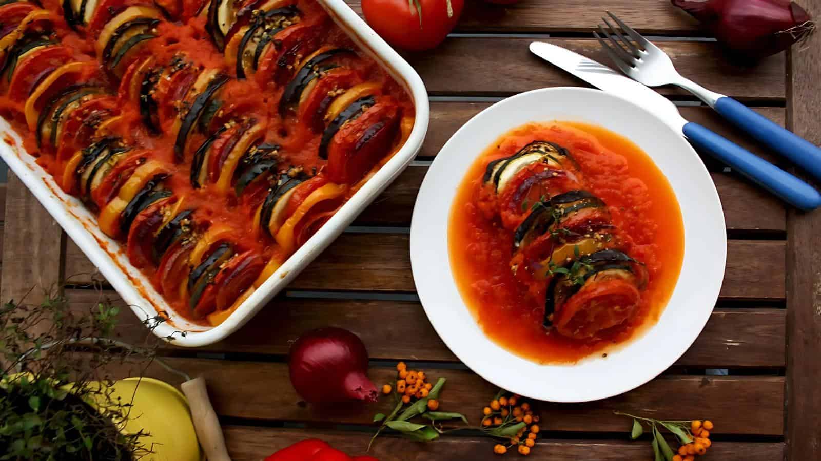A plate of ratatouille next to a baking dish filled with more of the dish, surrounded by vegetables and herbs on a wooden table.