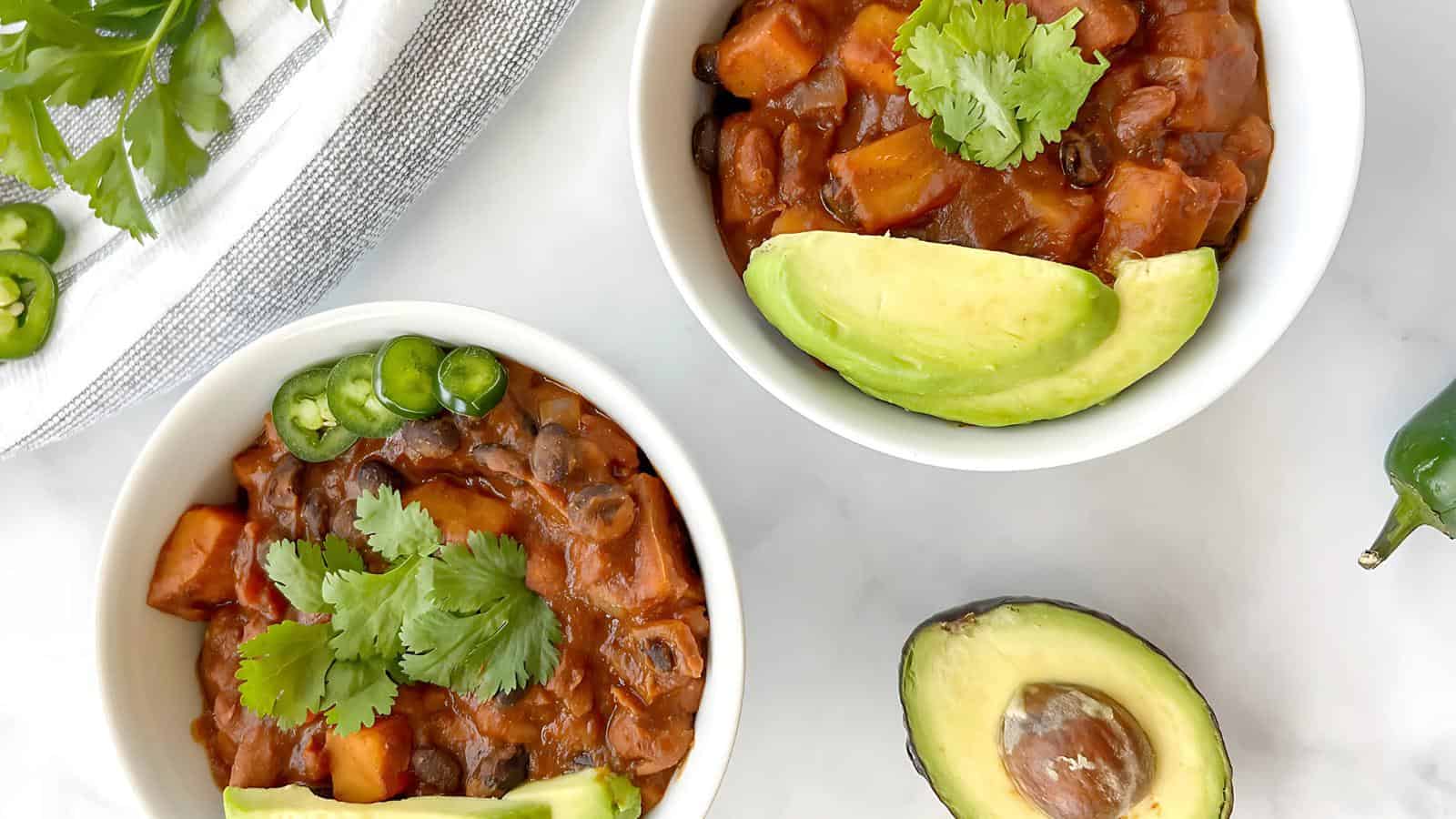 Two bowls of chili topped with cilantro, sliced jalapeños, and avocado slices sit on a table. An avocado half and sprigs of cilantro are nearby.