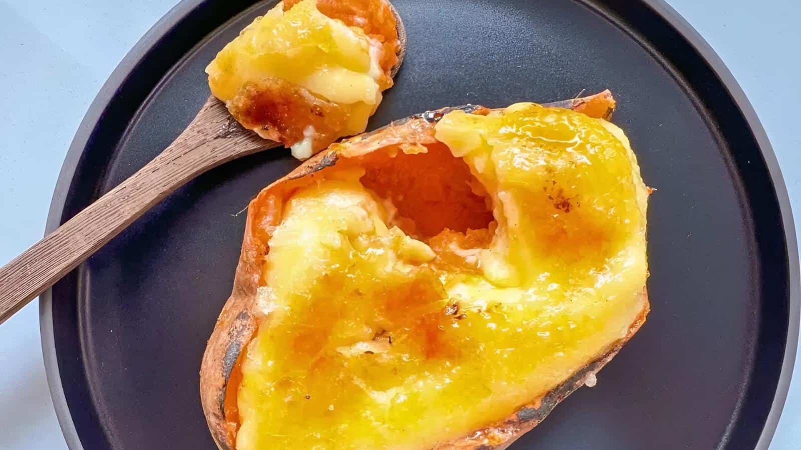 A baked sweet potato with a golden, melted topping on a black plate, accompanied by a wooden spoon.