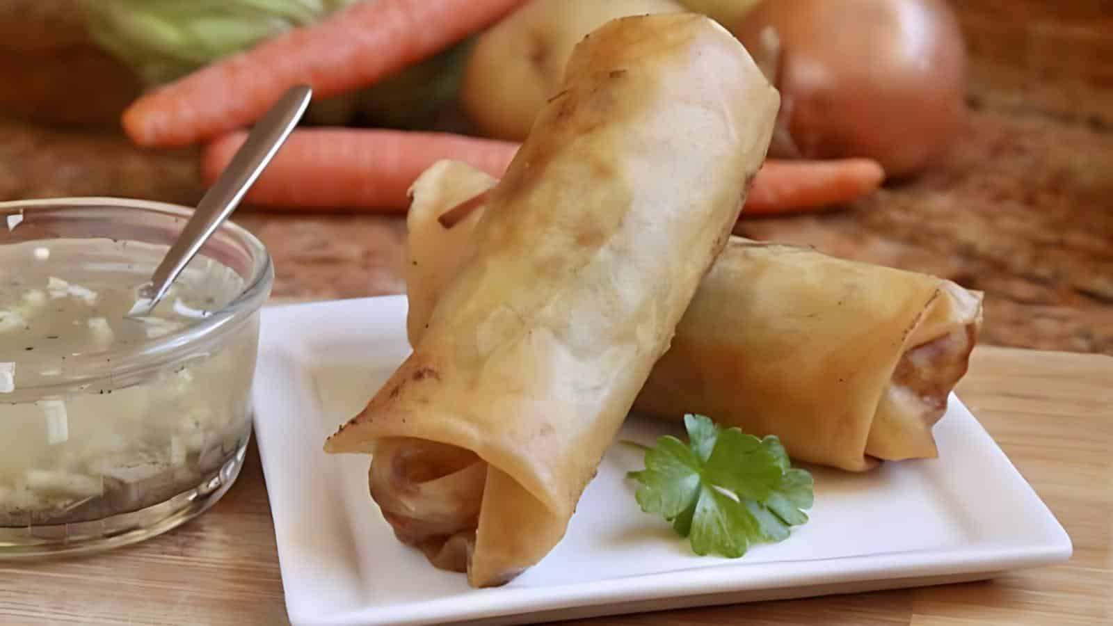 Two golden-brown spring rolls on a white plate with a parsley garnish, next to a bowl of dipping sauce. Vegetables like carrots and cabbage are blurred in the background.