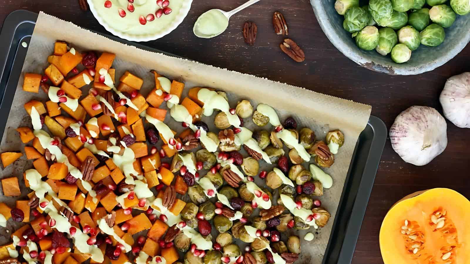 Roasted vegetables on a baking sheet, topped with white sauce and pomegranate seeds. Brussels sprouts, pecans, and squash cubes are visible. Garlic bulbs and a bowl are on the side.