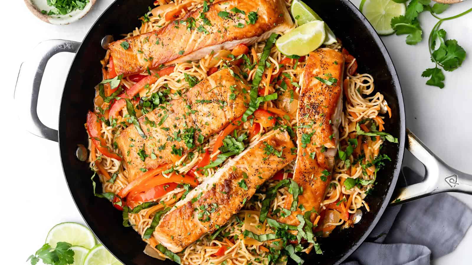 A skillet with cooked salmon fillets on a bed of stir-fried noodles and vegetables, garnished with herbs and lime wedges.