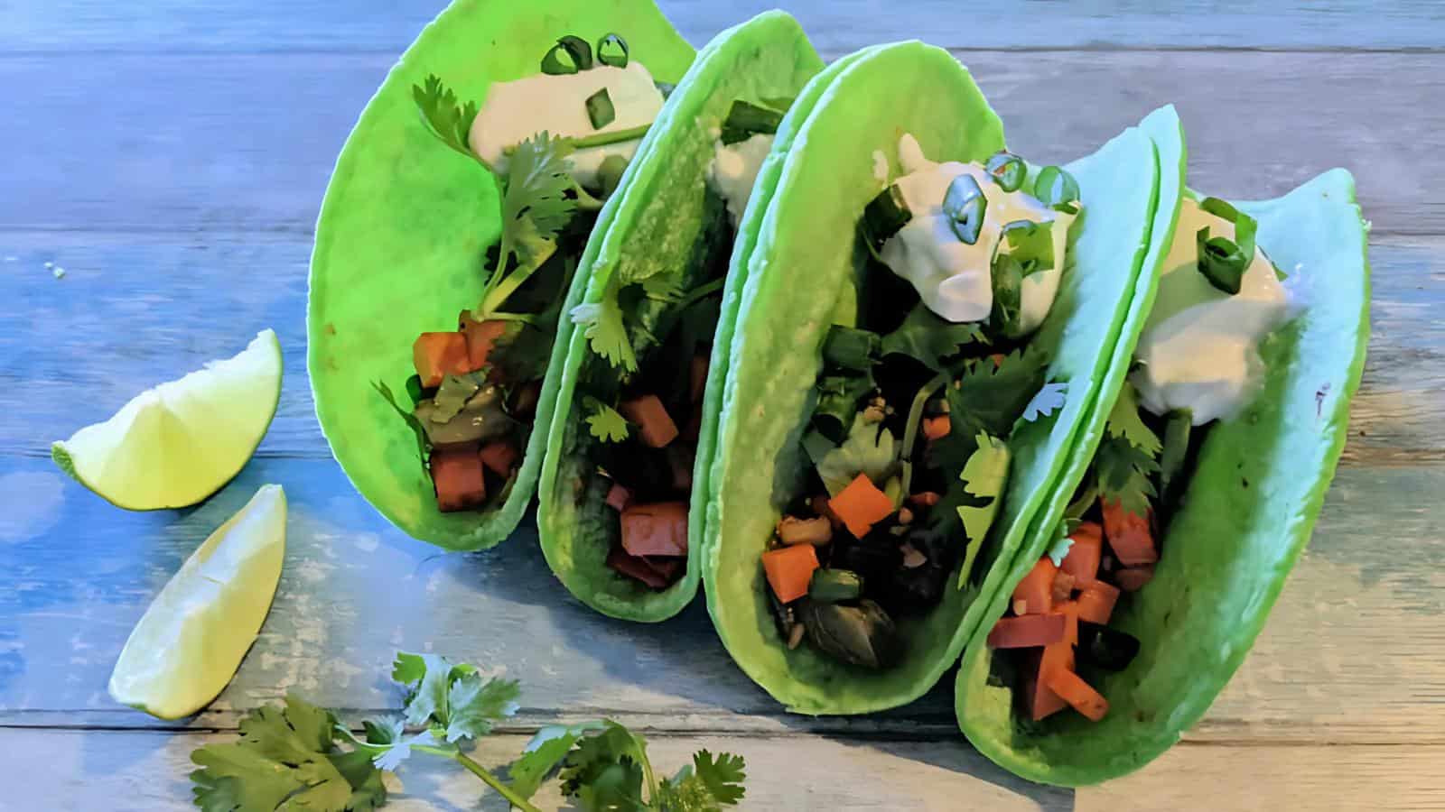 Four green tacos filled with diced vegetables, cream, and herbs on a wooden surface. Lime wedges and cilantro garnish are on the side.