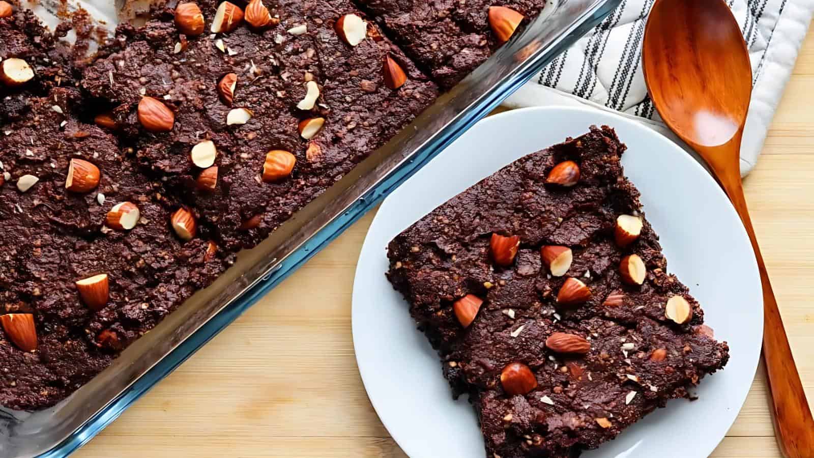 Chocolate brownies with almonds in a glass baking dish and on a white plate beside a wooden spoon on a striped cloth.