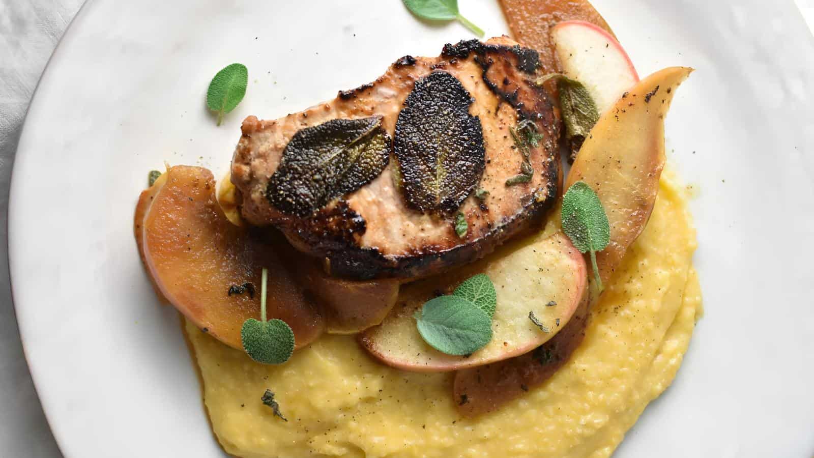 A plate with grilled pork chop topped with herbs, served over a bed of apple slices and creamy polenta.