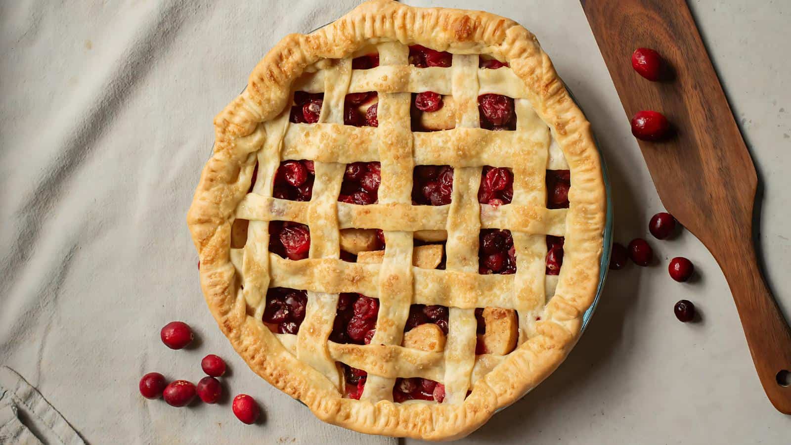 A lattice-top cranberry pie on a table with scattered cranberries and a wooden cutting board beside it.