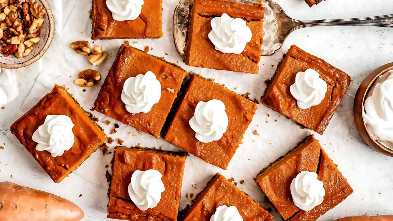 Pumpkin bars topped with dollops of whipped cream arranged on a white surface, with a serving spatula and walnuts beside them.