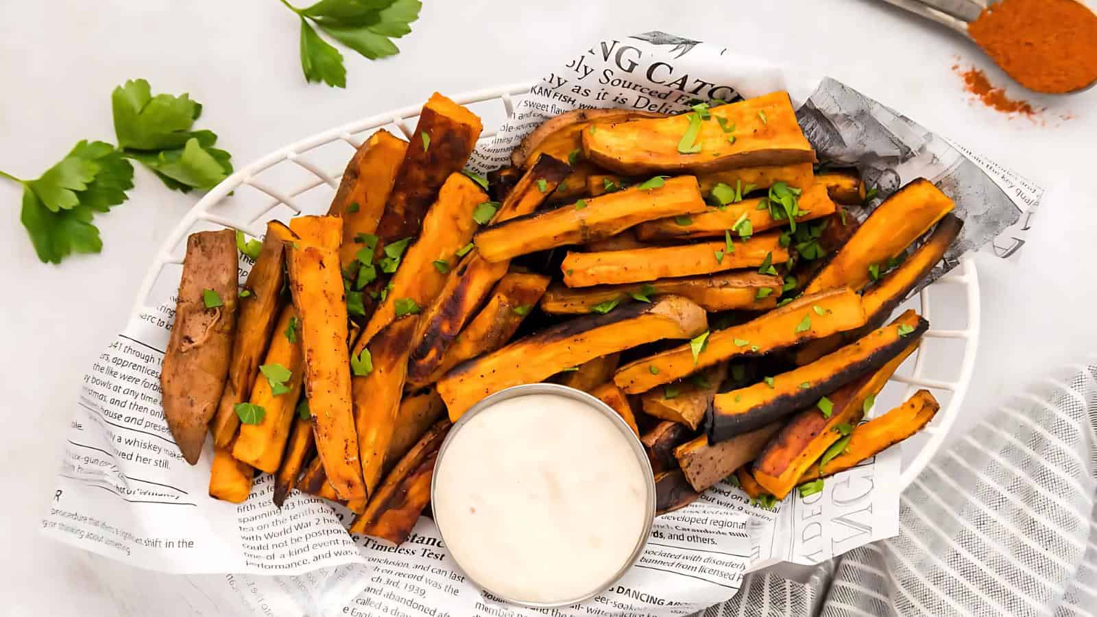 A plate of sweet potato fries garnished with parsley, served with a dipping sauce, placed on a newspaper-lined basket. Fresh parsley and a spoon with seasoning are in the background.
