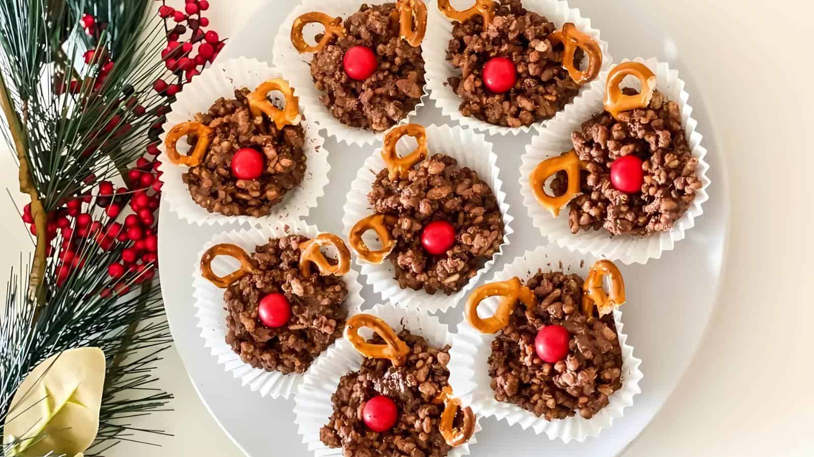 Plate of reindeer-themed chocolate treats with pretzel antlers and red candy noses, surrounded by holiday decorations.