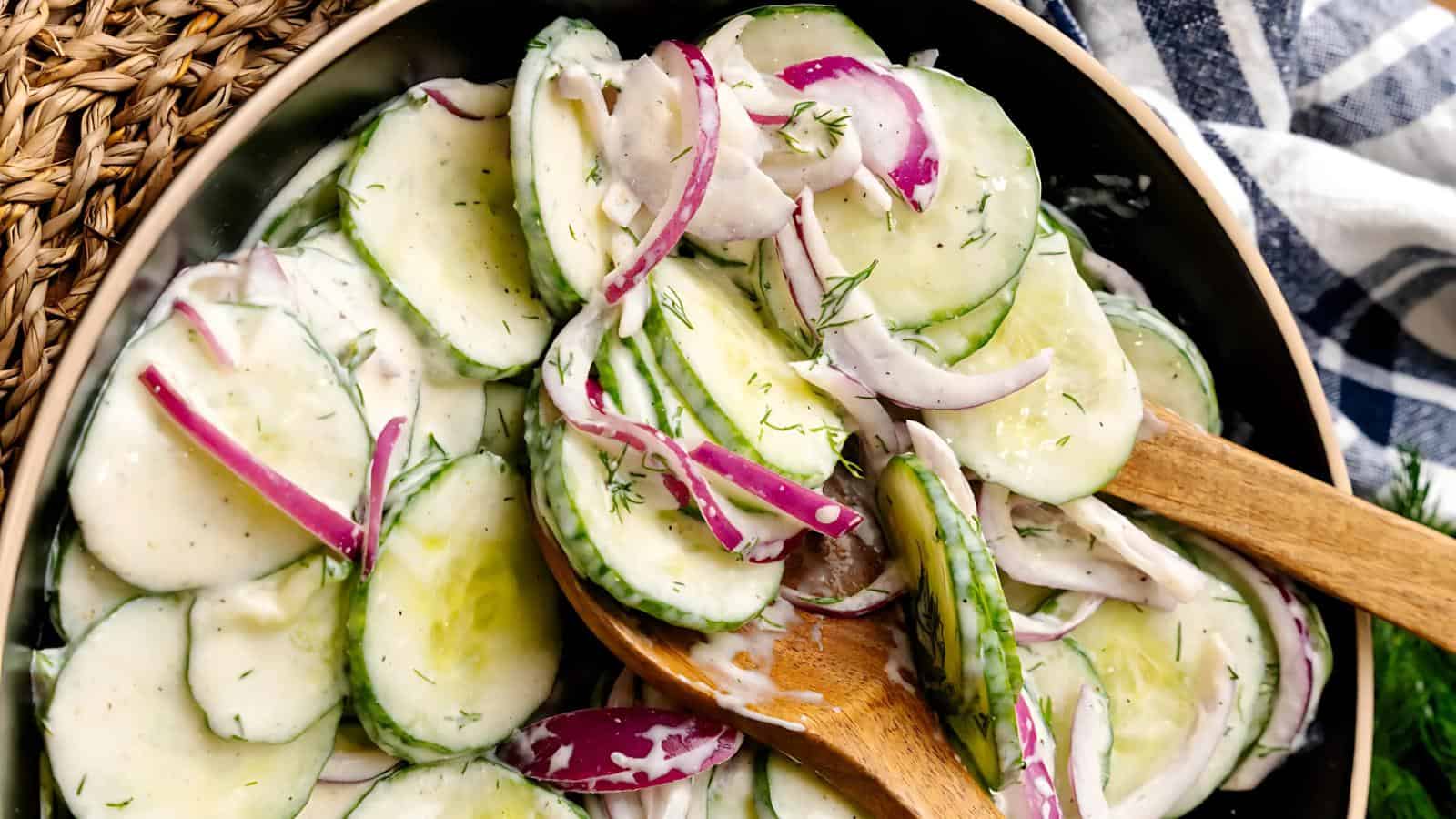 A bowl of cucumber and red onion salad mixed with creamy dressing, garnished with dill, with wooden salad tongs.