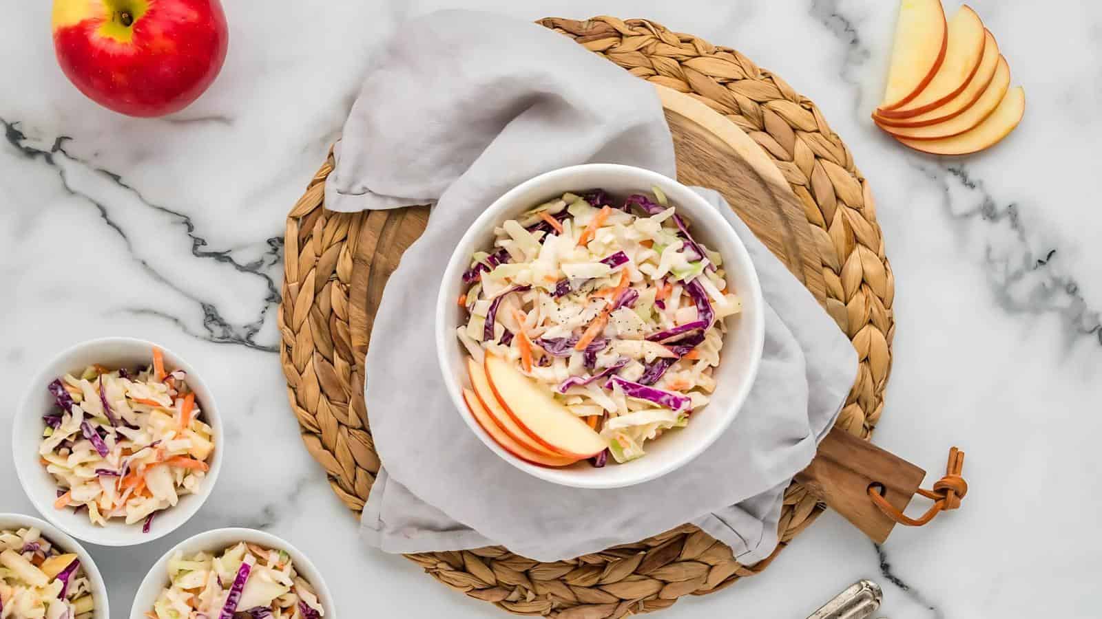 A bowl of coleslaw with apple slices on a woven mat, surrounded by additional bowls of coleslaw, an apple, and sliced apples on a marble surface.