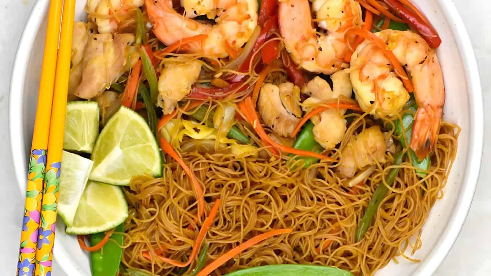 A plate of stir-fried noodles with shrimp, chicken, vegetables, and lime wedges, accompanied by colorful chopsticks.
