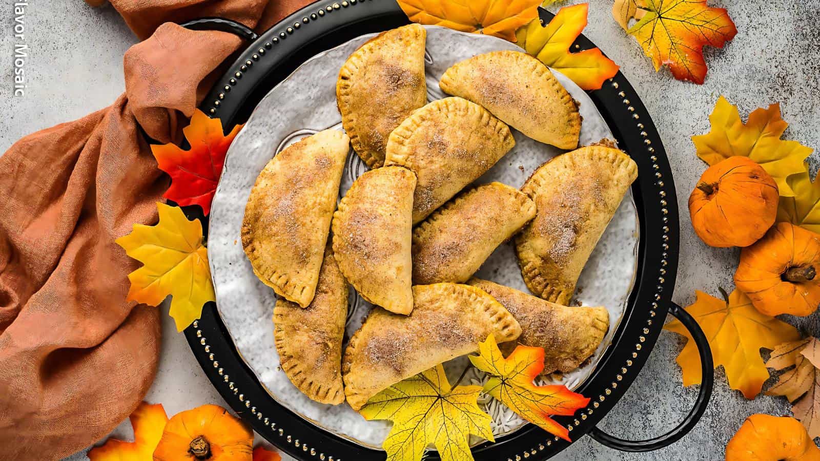 A plate of baked hand pies surrounded by orange and yellow autumn leaves and mini pumpkins.