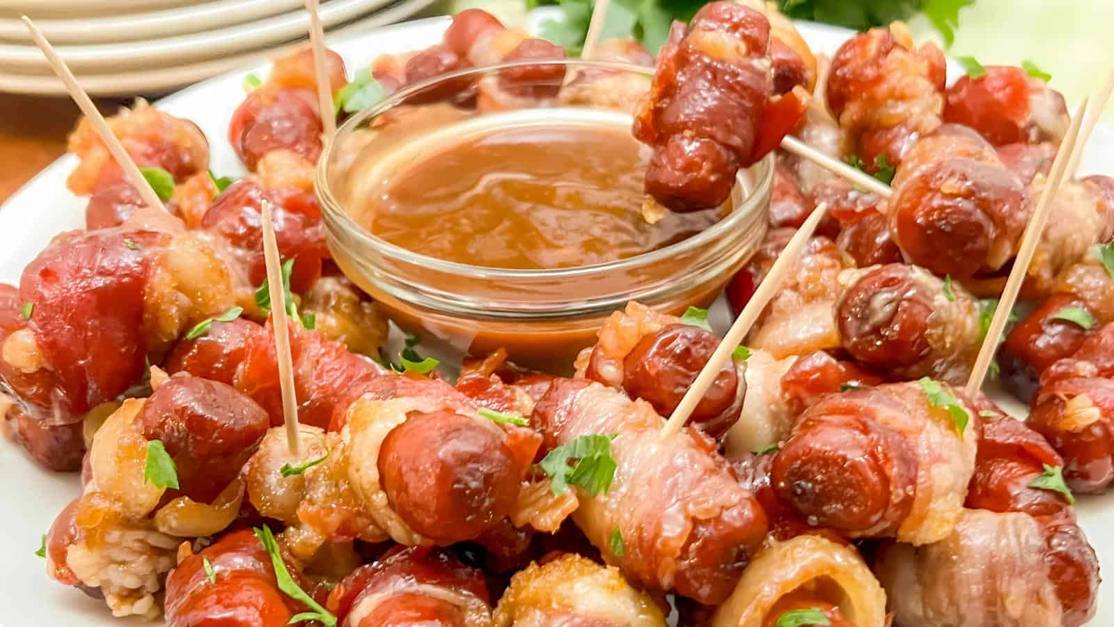 A plate of bacon-wrapped sausages with toothpicks, surrounding a bowl of dipping sauce.