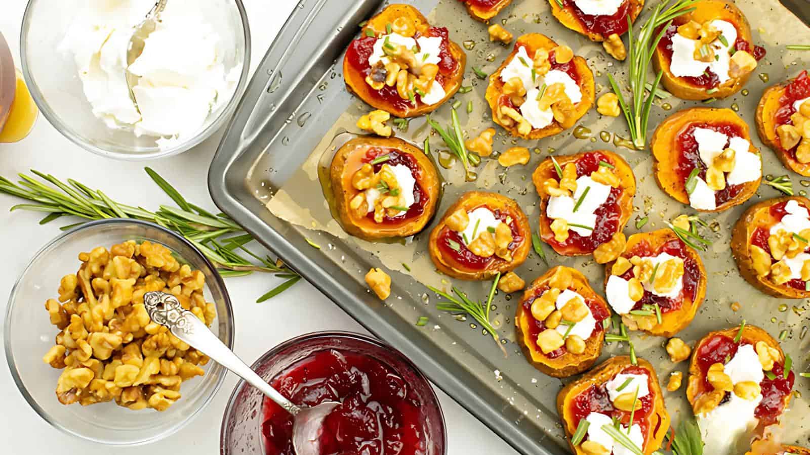 A baking tray with roasted sweet potato rounds topped with cheese, walnuts, and cranberry sauce. Bowls of walnuts, rosemary, cheese, and cranberry sauce are nearby.