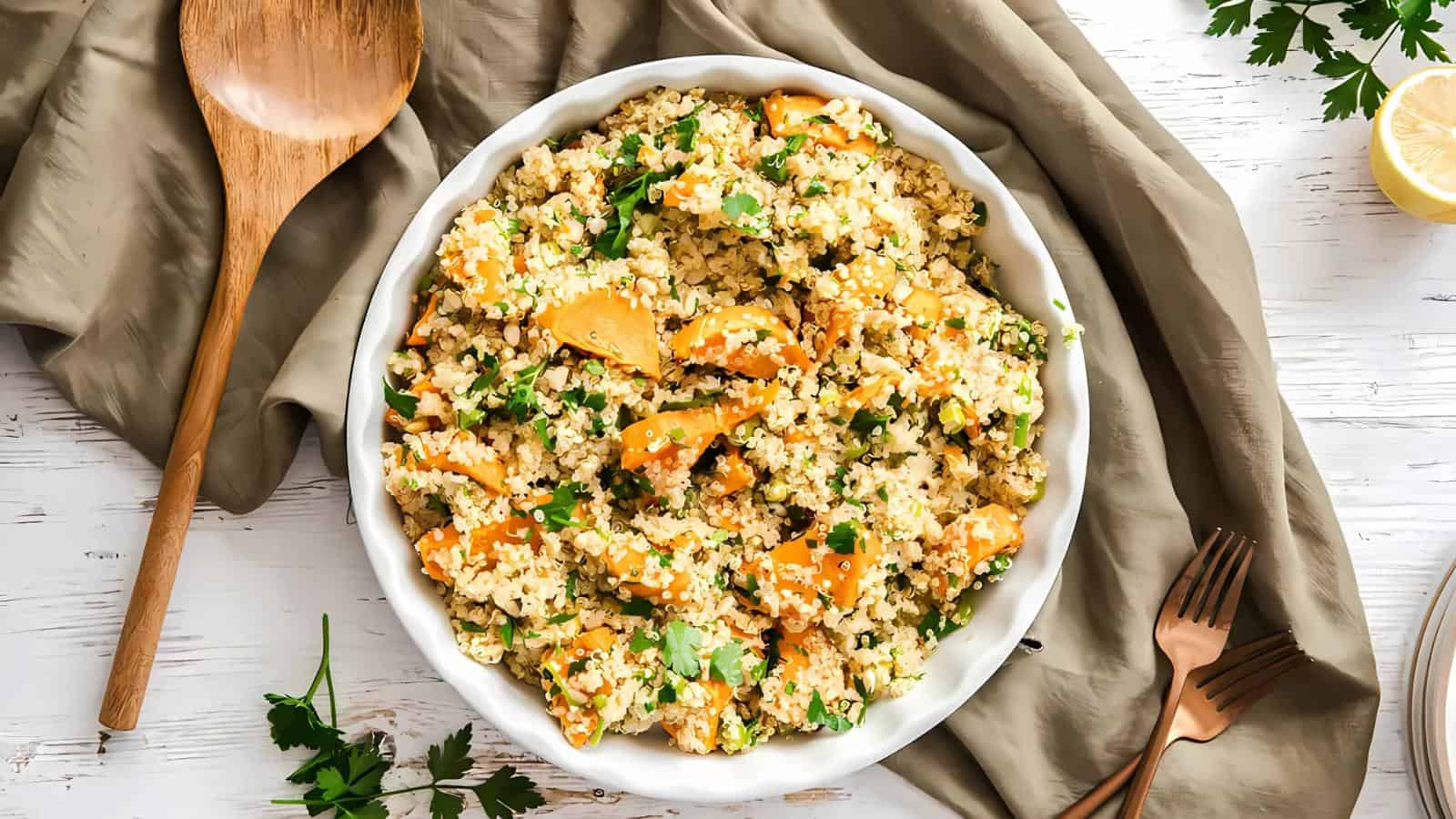 A bowl of quinoa salad with roasted butternut squash and fresh herbs, placed on a gray cloth. A wooden spoon and gold cutlery are nearby.