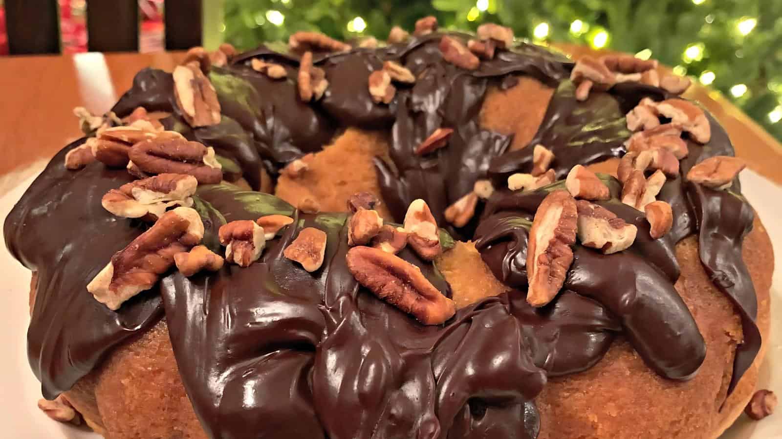 A bundt cake topped with glossy chocolate icing and chopped pecans, set against a background of blurred lights.
